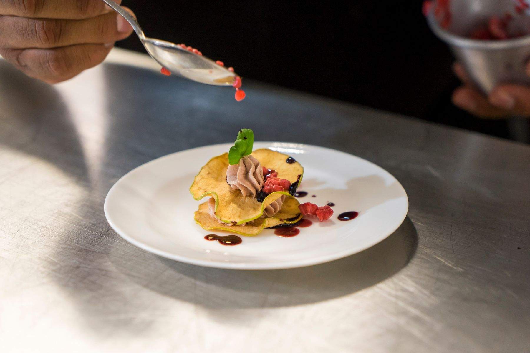 A chef spooning sauce onto a plated dish on a stainless-steel kitchen pass